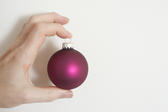 Man holding a colorful purple Christmas bauble in his fingers displaying it to the camera over a white background with copyspace for your holiday greeting